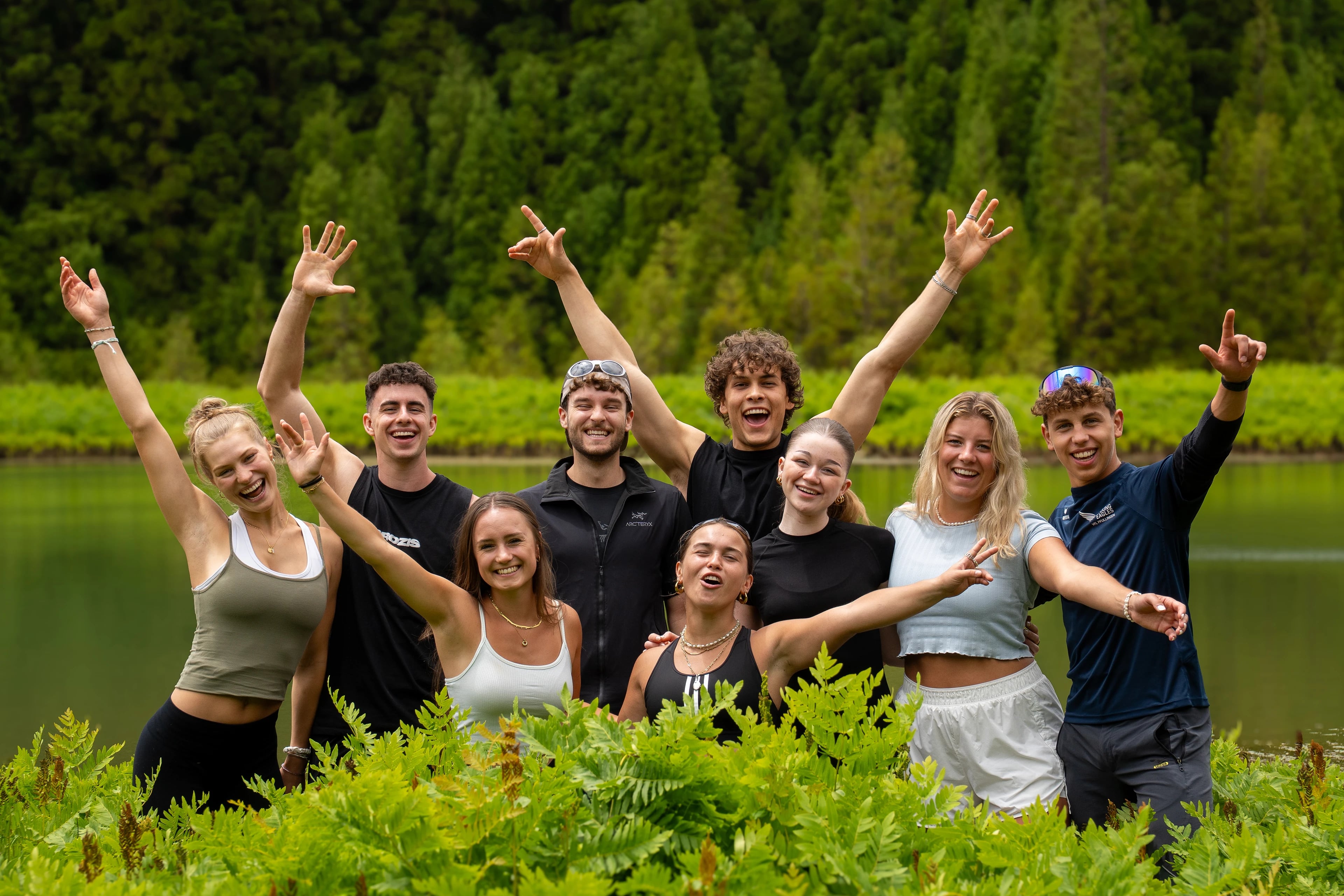 Eine MOHANA Gruppe jubelt gemeinsam mit erhobenen Armen am Ufer eines Kratersees auf den Azoren, dahinter dichter Wald.